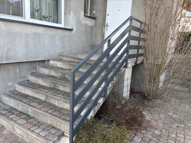 Zewnętrzne schody z szarymi, poziomymi poręczami metalowymi, prowadzące do białych drzwi wejściowych, otoczone surowym betonem i roślinnością.