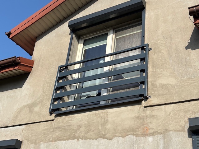 Szara, metalowa balustrada okienna z poziomymi szczebelkami zamontowana na elewacji budynku z widocznym oknem i roletą zewnętrzną.