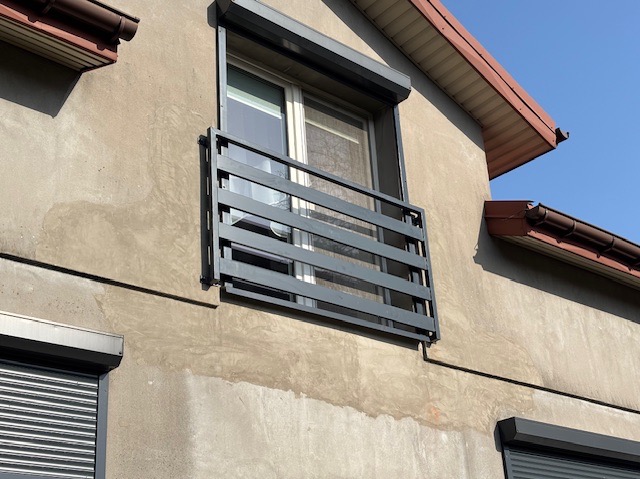 Szara, metalowa barierka okienna z poziomymi szczeblami zamontowana na elewacji budynku z roletą zewnętrzną.