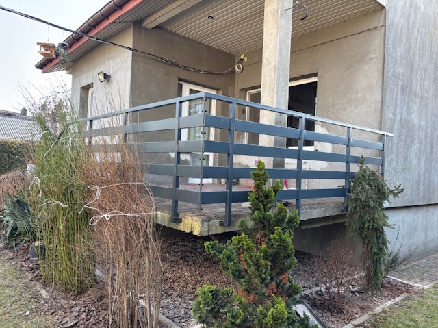 Szara, pozioma balustrada z metalowych profili na tarasie domu jednorodzinnego, widok z ogrodu, betonowa podstawa tarasu.