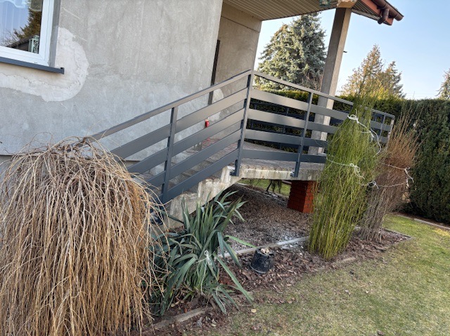 Zewnętrzne schody z metalową konstrukcją i poziomymi prętami, prowadzące do wejścia do domu, otoczone roślinnością.