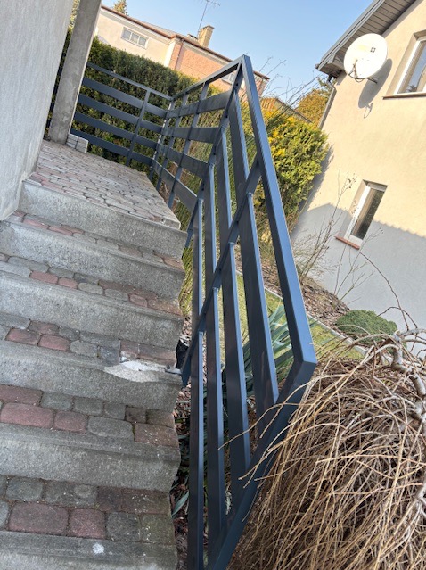 Szare metalowe zabezpieczenie schodów zewnętrznych z geometrycznym wzorem, widoczne z dołu, z elementami zieleni i budynków w tle.