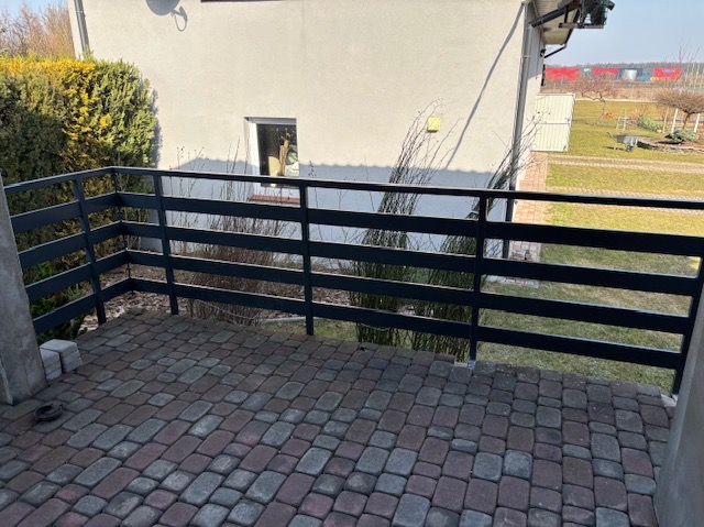 Czarna, pozioma balustrada z drewnianych desek na tarasie wyłożonym kostką brukową w odcieniach szarości i czerwieni. W tle jasny budynek z małym oknem i zielony krzew.