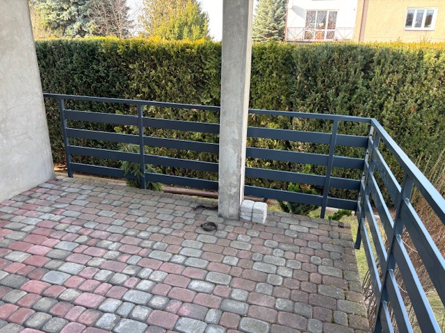 Ciemnoszara balustrada metalowa na tarasie z kostki brukowej w odcieniach czerwieni i szarości, z zielonym żywopłotem w tle.