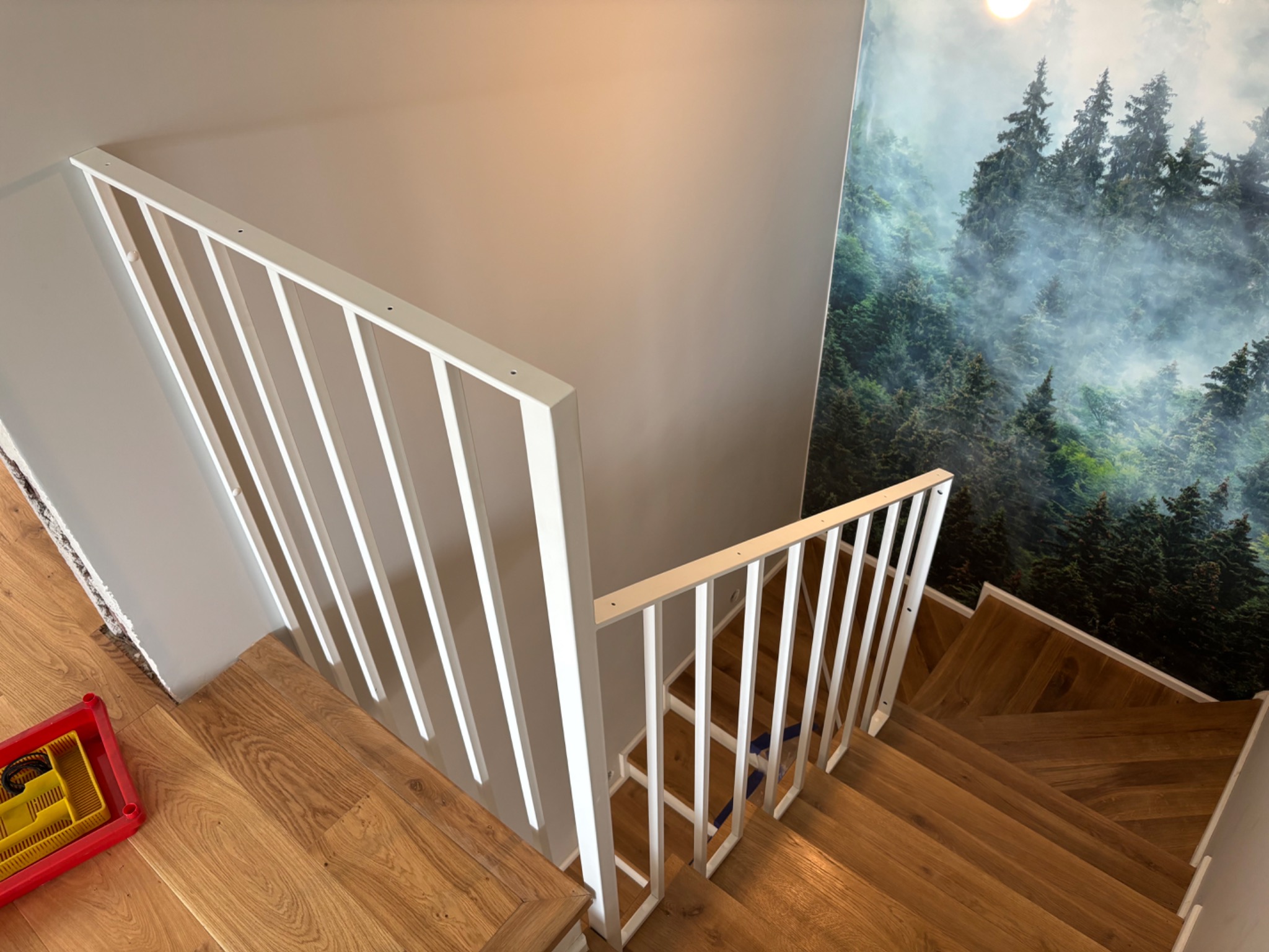 Nowoczesne, białe poręcze schodowe z pionowymi szczeblami, widok z góry na drewniane schody. W tle ściana z fototapetą leśną.