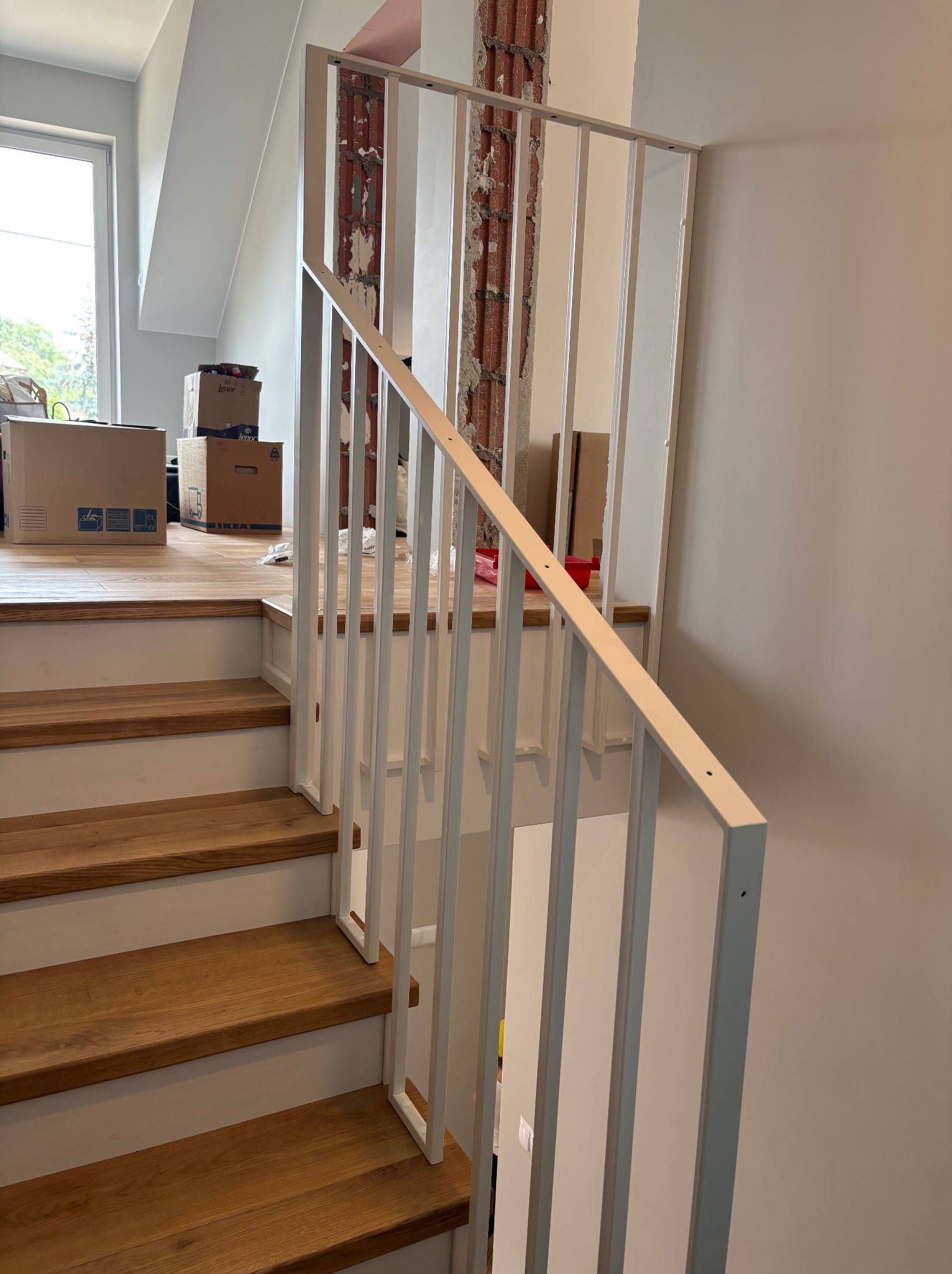 Biała, metalowa balustrada schodowa z drewnianymi stopniami w nowoczesnym wnętrzu. Widoczne elementy muru i kartony w tle. Ujęcie z dołu, perspektywa.