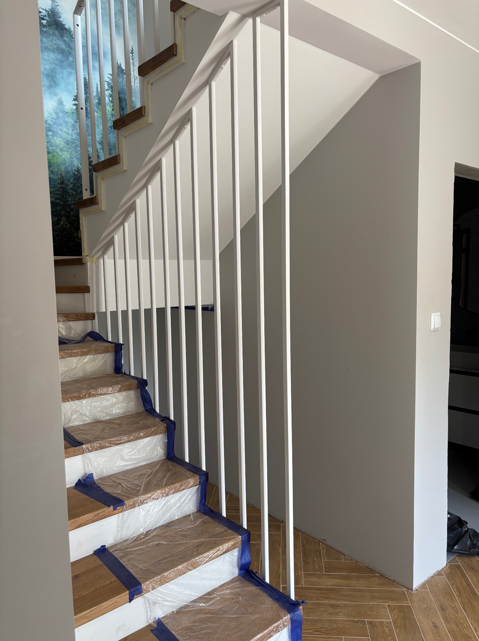 Biała, nowoczesna balustrada schodowa z pionowymi szczeblami. Schody drewniane zabezpieczone folią malarską. W tle fototapeta leśna i szara ściana.