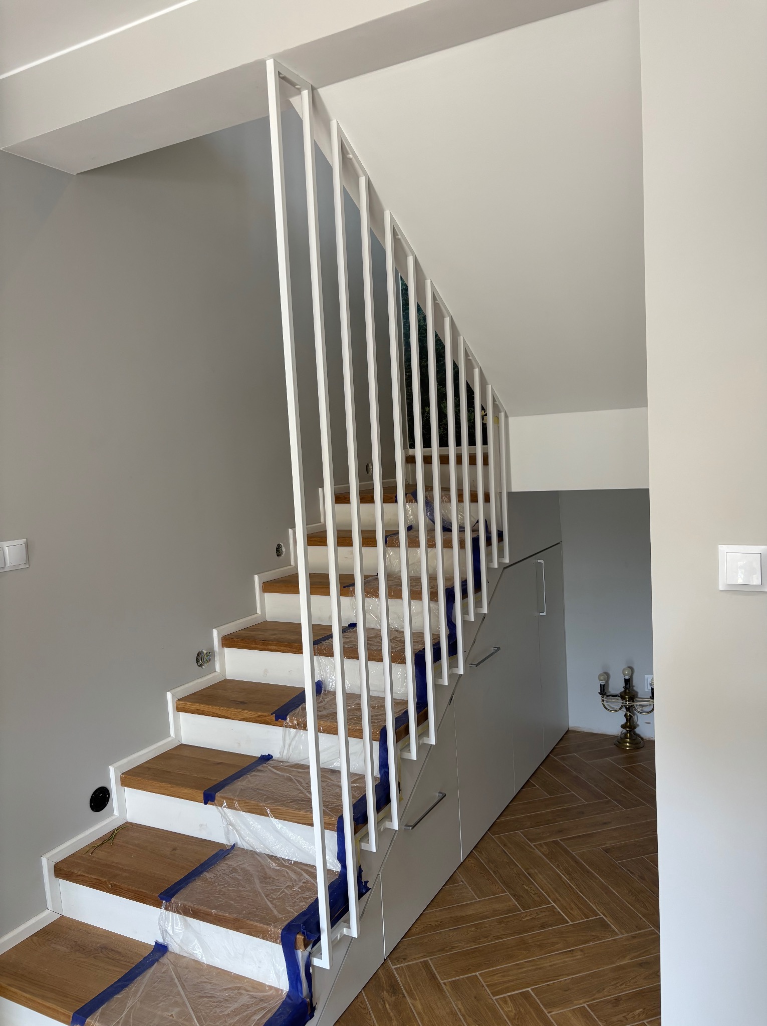 Minimalistyczne schody z drewnianymi stopniami i białą, pionową balustradą metalową. Stopnie zabezpieczone folią. Pod schodami zabudowa meblowa.