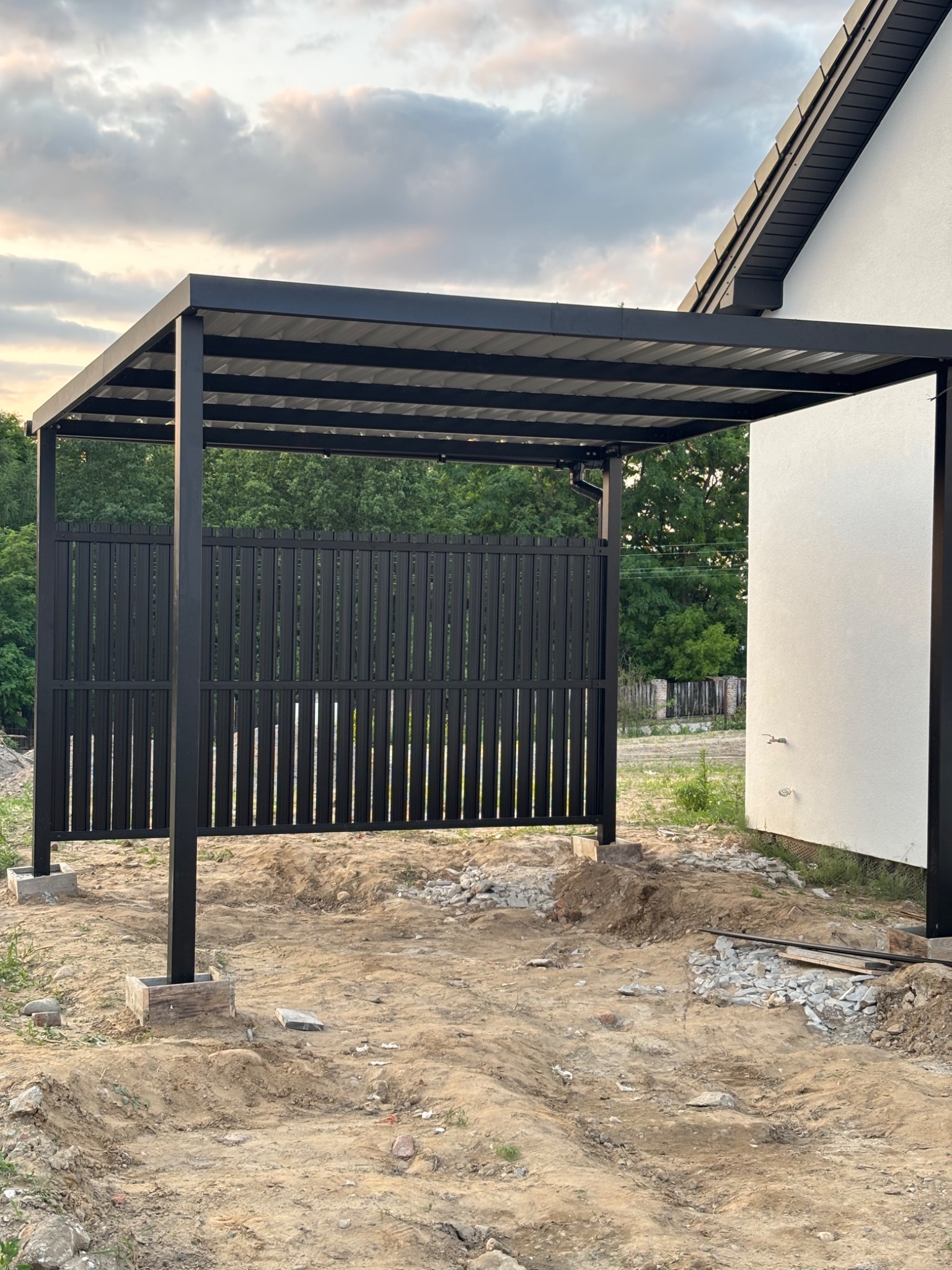 Nowoczesna, czarna konstrukcja zadaszenia z elementami ażurowymi na tle domu i zieleni, w trakcie montażu na nierównym terenie. Widoczne fundamenty słupów.
