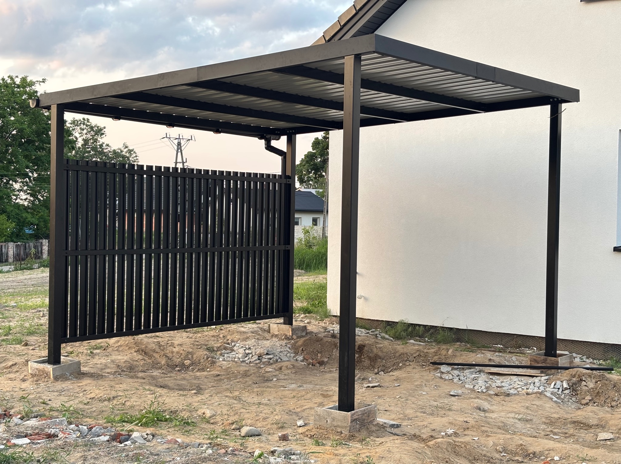 Nowoczesna, czarna konstrukcja zadaszenia z metalu z pionowymi panelami ogrodzeniowymi, osadzona na betonowych fundamentach. Widok na dom w tle.