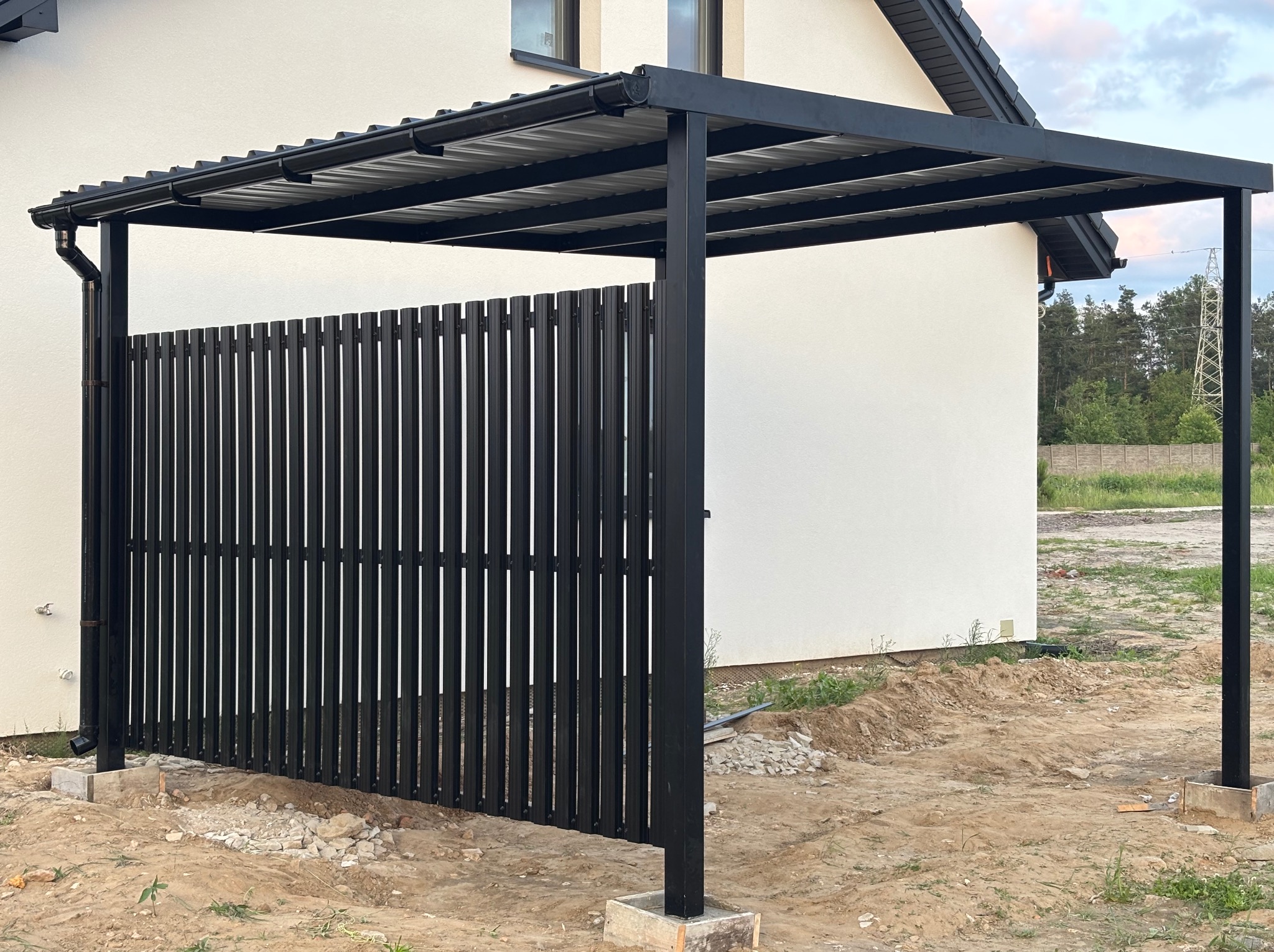Nowoczesna, czarna metalowa konstrukcja z zadaszeniem i pionowymi panelami ogrodzeniowymi na betonowych podstawach, na tle białego budynku i zieleni.