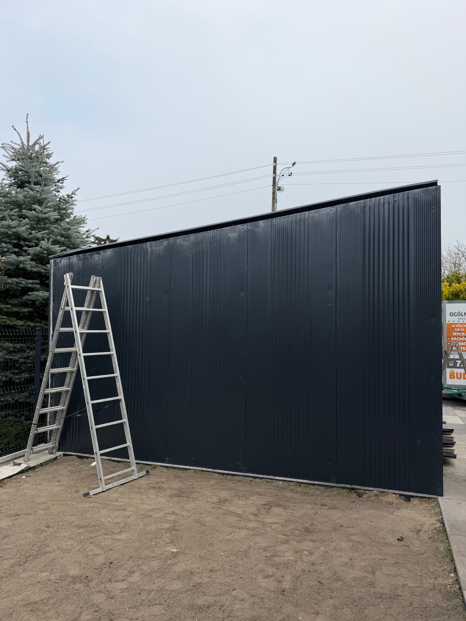 Ciemna, metalowa brama przemysłowa z drabinką opartą o bok, na tle pochmurnego nieba i zieleni. Widoczne pionowe panele z delikatnym żłobieniem.