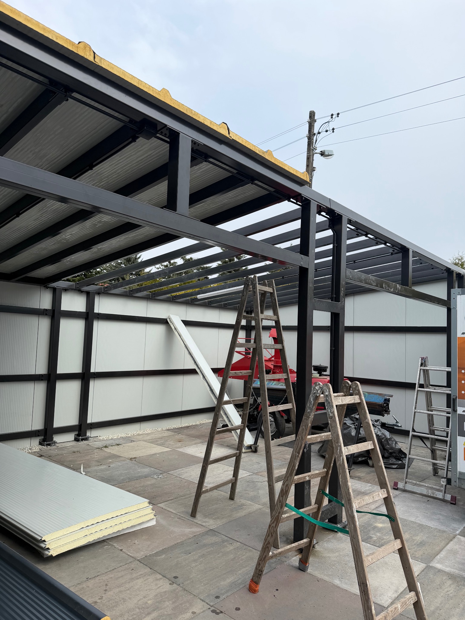 Konstrukcja z czarnych profili stalowych w trakcie montażu z użyciem drabin. Widoczne płyty warstwowe i narzędzia. Ujęcie z dołu, na zewnątrz.