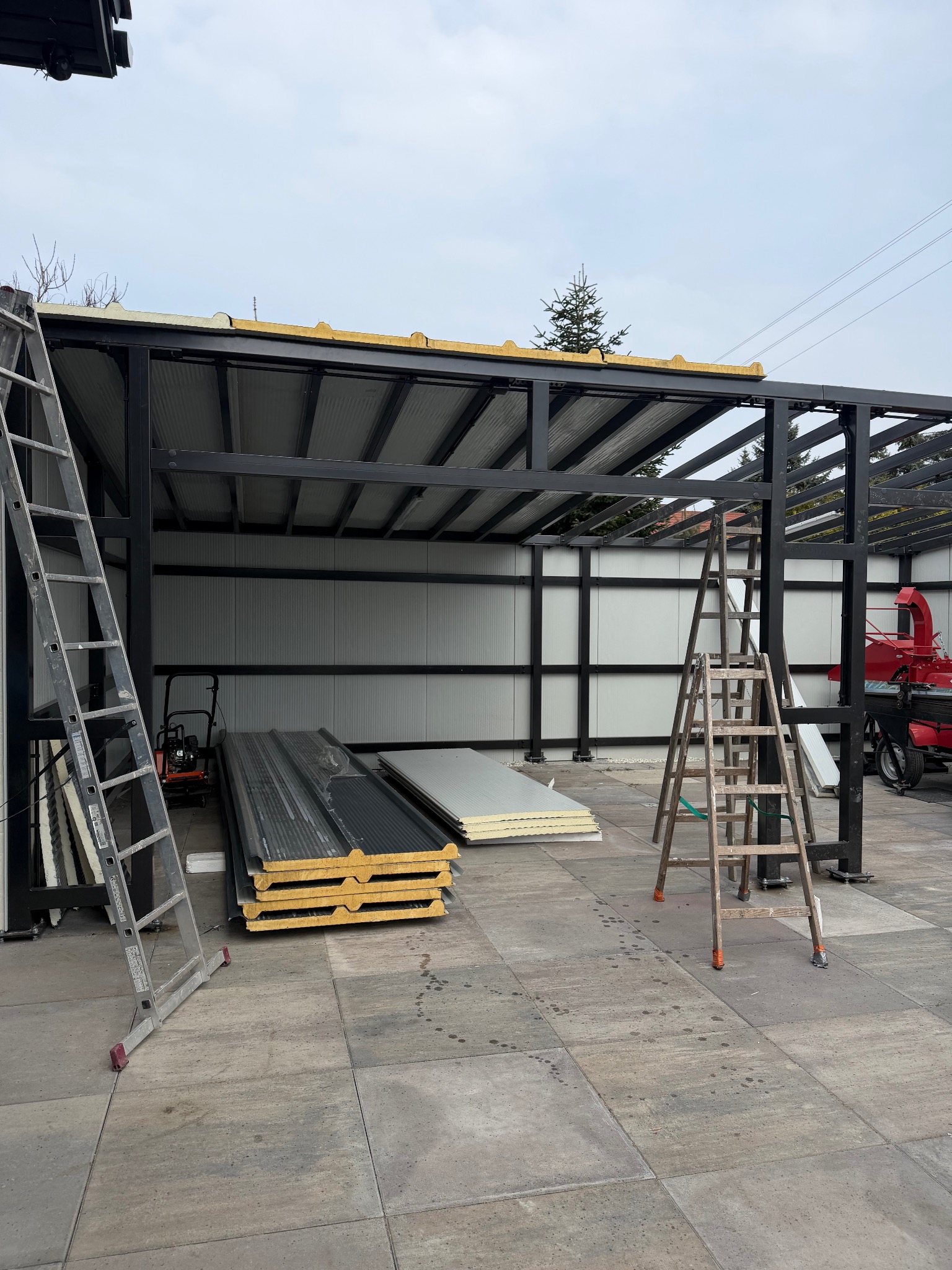 Konstrukcja z metalu w trakcie montażu, widoczne płyty warstwowe, drabiny i narzędzia. Elementy konstrukcyjne pomalowane na czarno. Tło stanowi szara kostka brukowa.