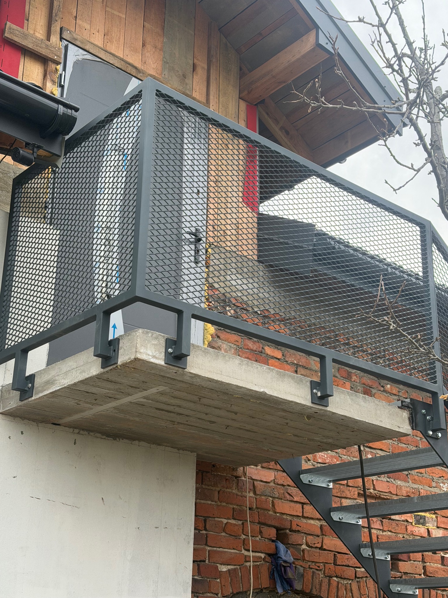 Zewnętrzna, metalowa konstrukcja balkonu z siatkową balustradą, zamocowana do betonowej płyty i ceglanej ściany budynku. Widoczne metalowe schody i fragment drzewa.