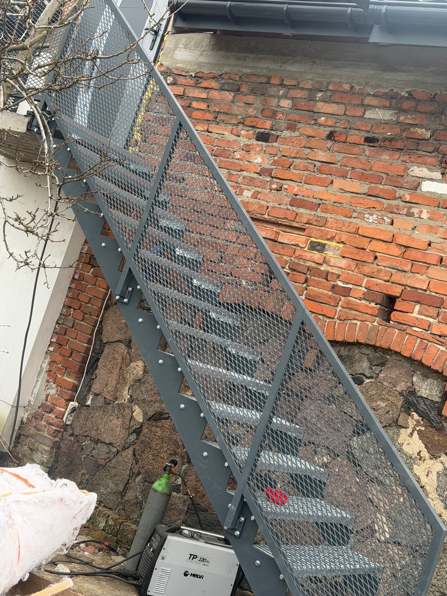 Zewnętrzne schody metalowe z siatkową osłoną, przymocowane do ceglano-kamiennej ściany budynku. Widoczny sprzęt spawalniczy u podnóża schodów.