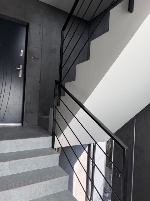 Nowoczesne schody z czarną, metalową balustradą o minimalistycznym designie. Szare płytki schodowe i kontrastujące, białe ściany. Widok z dołu, blisko drzwi wejściowych.