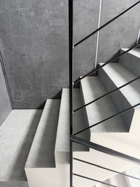 Minimalistyczne schody z szarymi stopniami i czarną, metalową barierką. Surowy beton na ścianie. Ujęcie z góry, podkreślające geometrię i nowoczesny design.