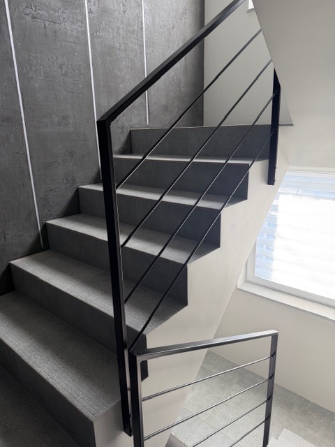 Nowoczesne schody z czarną, metalową konstrukcją i szarymi stopniami, oświetlone naturalnym światłem z okna. Minimalistyczny design z akcentem na geometrię.