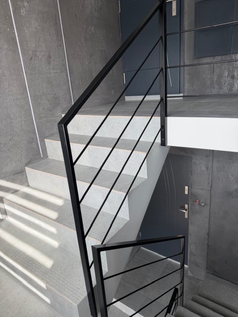 Minimalistyczne schody z czarną, metalową poręczą w nowoczesnym budynku. Widoczne drzwi na podeście i gra świateł na stopniach. Ujęcie z dołu.