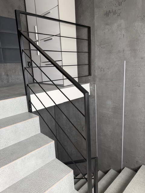 Minimalistyczna, czarna poręcz schodowa z poziomymi prętami na tle szarej ściany o fakturze betonu i białych szafek. Ujęcie z góry, widoczne stopnie schodów.