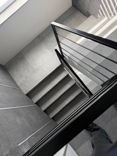 Minimalistyczne schody z czarną, metalową poręczą z prętów, widziane z góry. Szare płytki na stopniach i ścianach. Wpadające światło słoneczne tworzy wzory cieni.