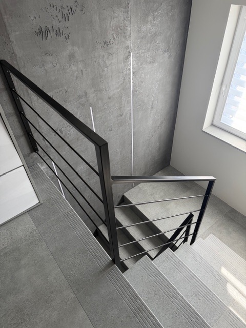 Minimalistyczne schody z czarną, metalową konstrukcją i poziomymi prętami, widok z góry, szare płytki, nowoczesny design, oświetlenie naturalne z okna.