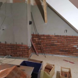 JKM Renovation Jakub Mrzygłód - Fragment ściany z cegły klinkierowej, w trakcie montażu, widoczne profile montażowe, skrzynki z cegłami, surowy stan ścian i poddasza.