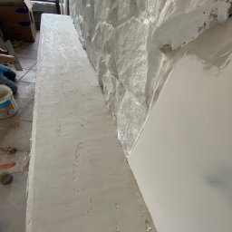 JKM Renovation Jakub Mrzygłód - Nakładanie białej gładzi na ścianę z kamienia, widoczne narzędzia i wiadro z farbą, fragment okna w tle.