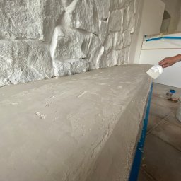 JKM Renovation Jakub Mrzygłód - Nakładanie szpachli na półkę pod ścianą z dekoracyjną imitacją kamienia, widoczny fragment ręki z narzędziem, wiadro z masą i zabezpieczone taśmą malarską krawędzie.