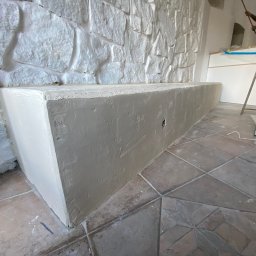 JKM Renovation Jakub Mrzygłód - Świeżo otynkowana, długa, narożna ławka z kamienną ścianą w tle, na podłodze terakota, w tle widoczna drabina i osoba pracująca przy suficie.