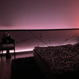 JKM Renovation Jakub Mrzygłód - Poddasze sypialni z niską ścianką działową, oświetlone taśmą LED w kolorze różowym, z dekoracyjną ścianą w geometryczne wzory, stolikiem nocnym z pluszową pandą i łóżkiem z pościelą w zebrę.