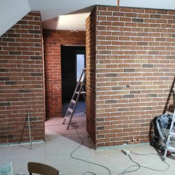 JKM Renovation Jakub Mrzygłód - Wnętrze poddasza z wykończonymi ścianami z cegły klinkierowej w odcieniach brązu i beżu, widoczne drabiny malarskie i narzędzia, gniazdka elektryczne w ścianie.