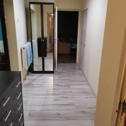 JKM Renovation Jakub Mrzygłód - Wnętrze mieszkania z nową podłogą imitującą drewno, widoczna szafa z lustrem, grzejnik z ręcznikiem i fragment komody.