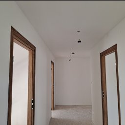 JKM Renovation Jakub Mrzygłód - Korytarz w trakcie remontu z zamontowanymi, drewnianymi ościeżnicami drzwiowymi i przygotowanymi punktami oświetleniowymi na suficie.
