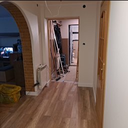 JKM Renovation Jakub Mrzygłód - Wnętrze domu w trakcie prac wykończeniowych: świeżo ułożona podłoga z paneli, widoczne narzędzia i materiały budowlane w przejściu, żółty worek z odpadami, grzejnik, oraz widoczny fragment...