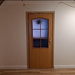 JKM Renovation Jakub Mrzygłód - Drewniane drzwi wewnętrzne z szybami o fakturze szkła ornamentowego, umieszczone w białej ścianie, widoczne z perspektywy łuku wejściowego z ceglaną okładziną.