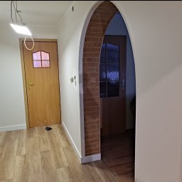 JKM Renovation Jakub Mrzygłód - Korytarz z drewnopodobną podłogą, dwoje drzwi z ościeżnicami, jedno z ceglanym łukiem, surowa instalacja oświetleniowa na suficie.