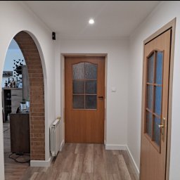 JKM Renovation Jakub Mrzygłód - Korytarz mieszkania po remoncie z dwiema drewnianymi drzwiami z szybami, łukowate wejście do pokoju z ceglanym obramowaniem, grzejnik i panele podłogowe imitujące drewno.