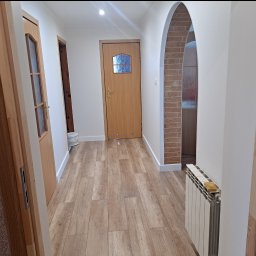 JKM Renovation Jakub Mrzygłód - Korytarz po remoncie z panelami podłogowymi imitującymi drewno, widoczne trzy drewniane drzwi, łuk z cegły i grzejnik z gąbką na górze.