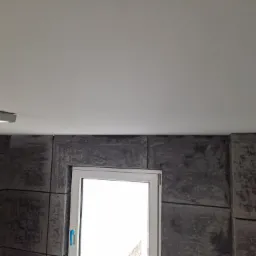 Szara ściana z efektem betonu architektonicznego, obramowana czarnymi liniami, białe okno z niebieskim zabezpieczeniem klamki, biały sufit.