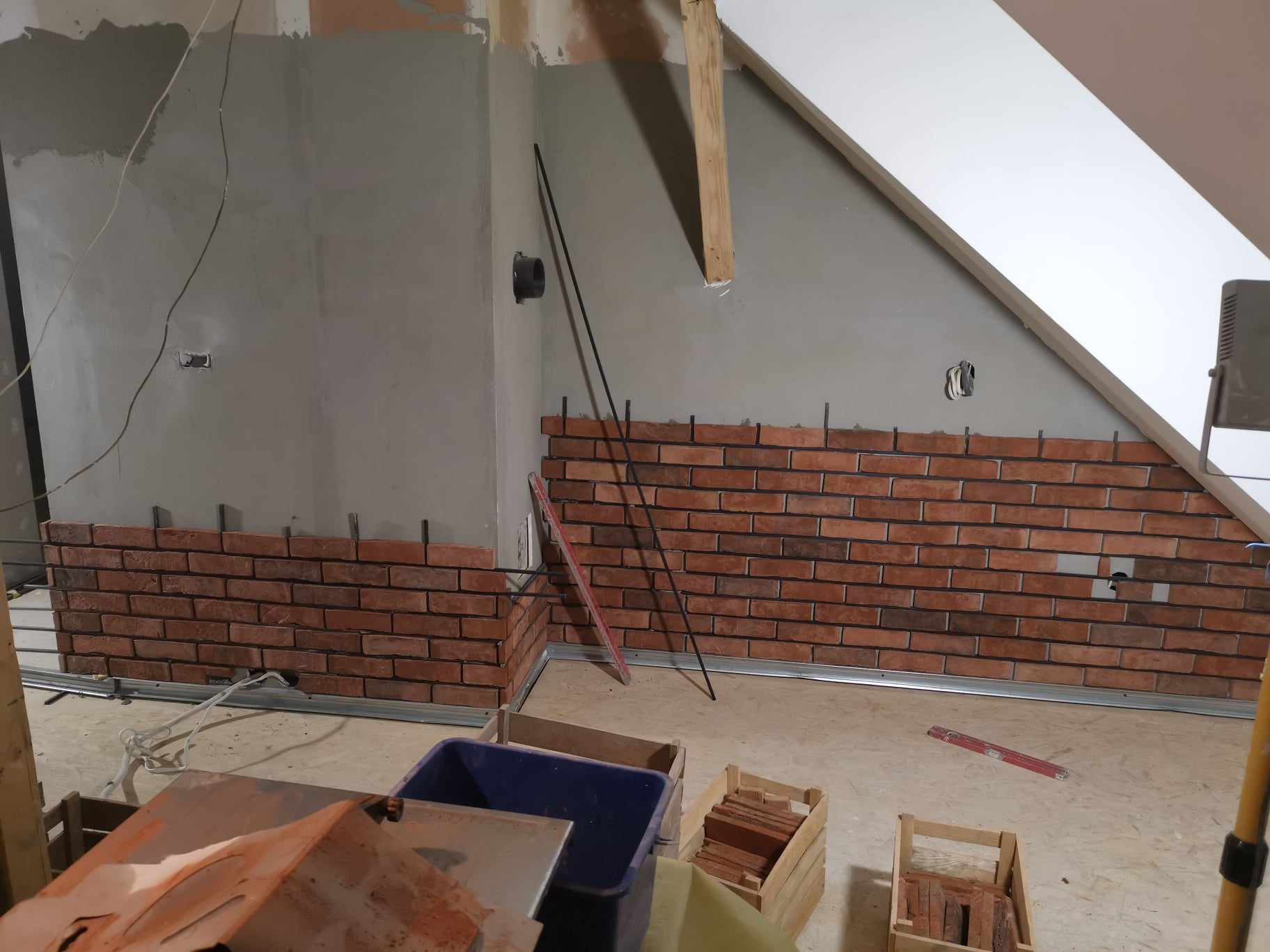 Wnętrze poddasza w trakcie wykańczania, fragment ściany z cegieł klinkierowych, widoczne profile montażowe, skrzynki z cegłami i narzędzia.