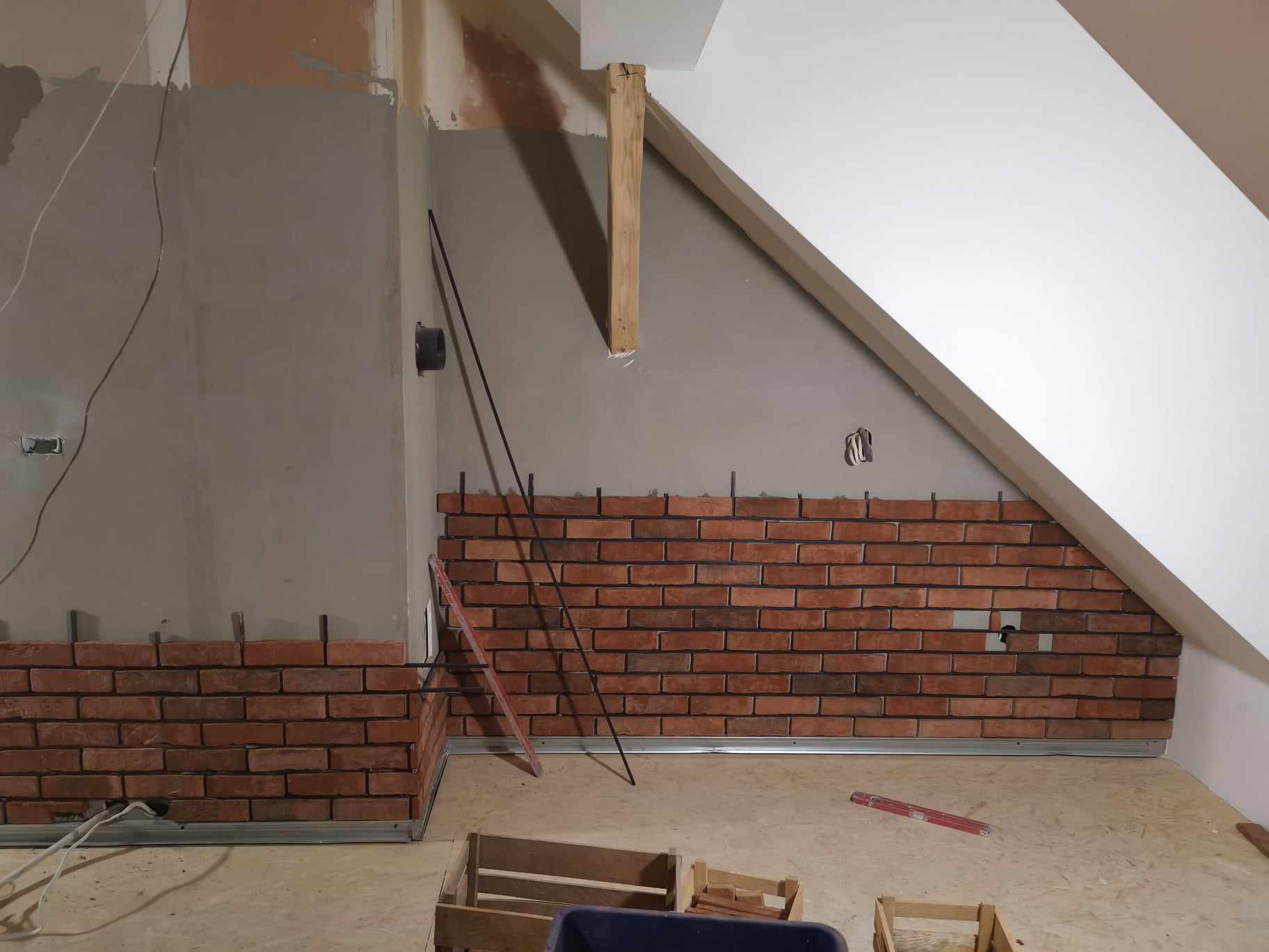 Montaż dekoracyjnej ściany z cegły w pomieszczeniu na poddaszu, widoczne elementy mocujące i narzędzia pracy.
