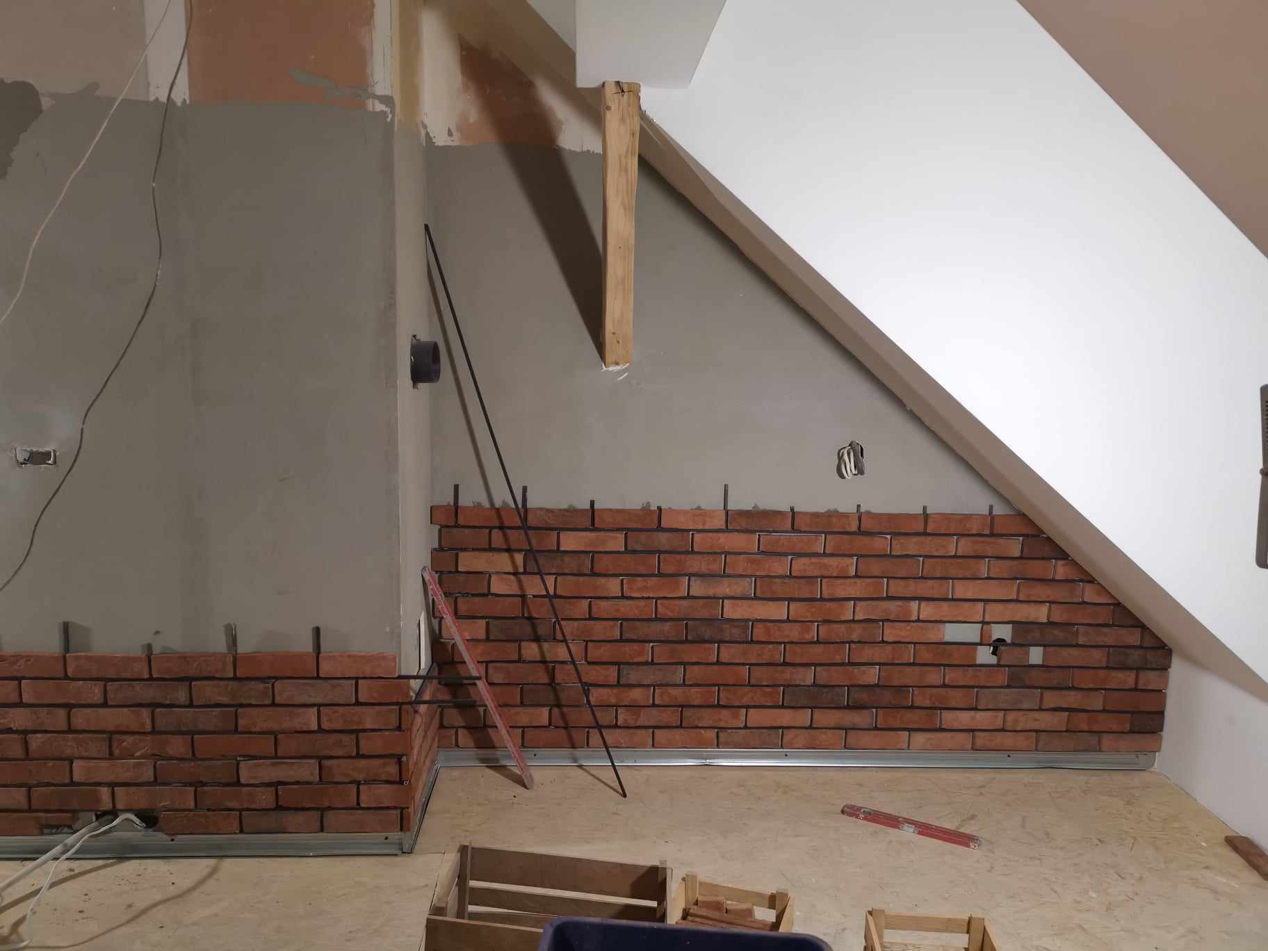 Montaż cegieł dekoracyjnych na ścianie pod skosem, widoczne pręty montażowe i poziomica, surowy stan wykończenia ścian i podłogi.