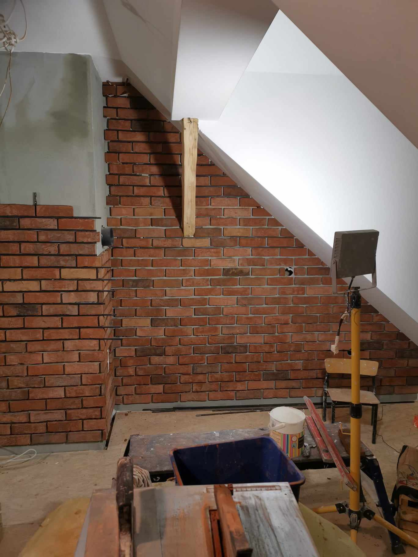 Ceglana ściana w trakcie budowy na poddaszu, widoczny fragment niedokończonej ściany, drewniany stempel podtrzymujący konstrukcję, narzędzia i materiały budowlane w tle.