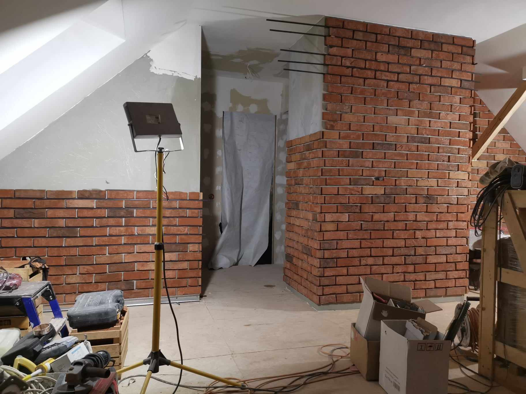 Montaż dekoracyjnej ściany z cegły w surowym wnętrzu poddasza, widoczny reflektor budowlany i narzędzia pracy.