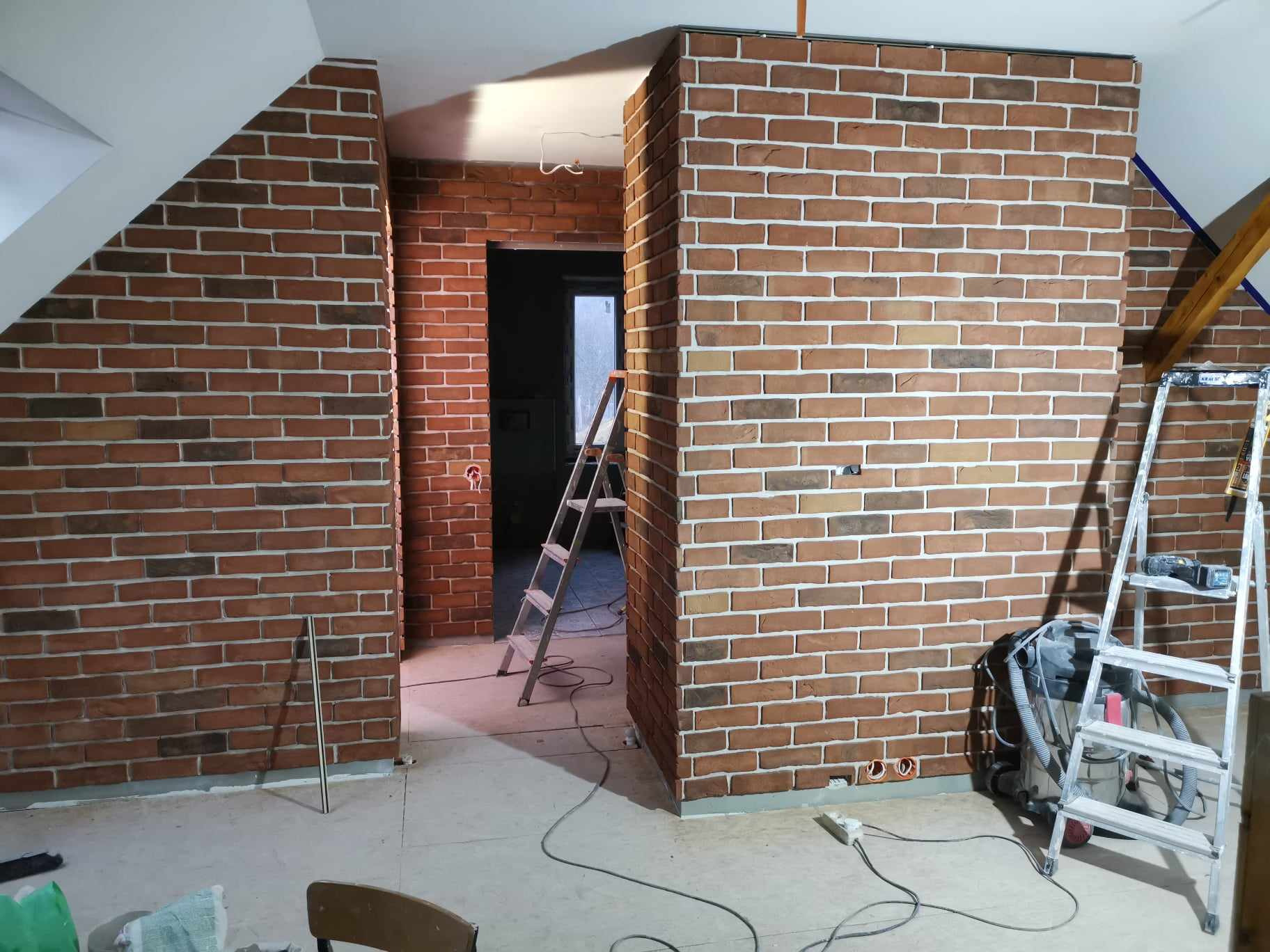Wnętrze poddasza z wykończonymi ścianami z cegły klinkierowej w odcieniach brązu i beżu, widoczne drabiny malarskie i narzędzia, gniazdka elektryczne w ścianie.