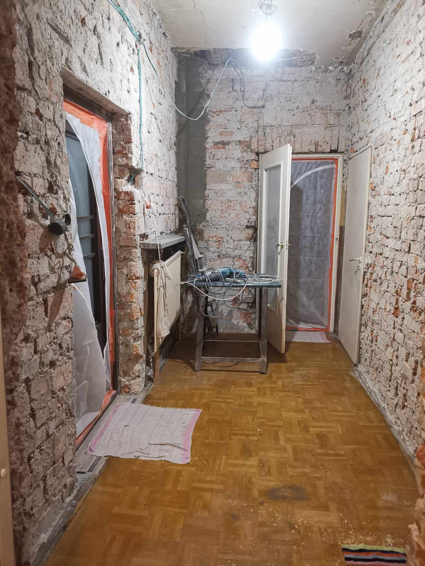 Wnętrze w trakcie rozbiórki: odsłonięta cegła na ścianach, zabezpieczone folią drzwi, drewniana podłoga, stolik z elektronarzędziem i wiszące przewody elektryczne.
