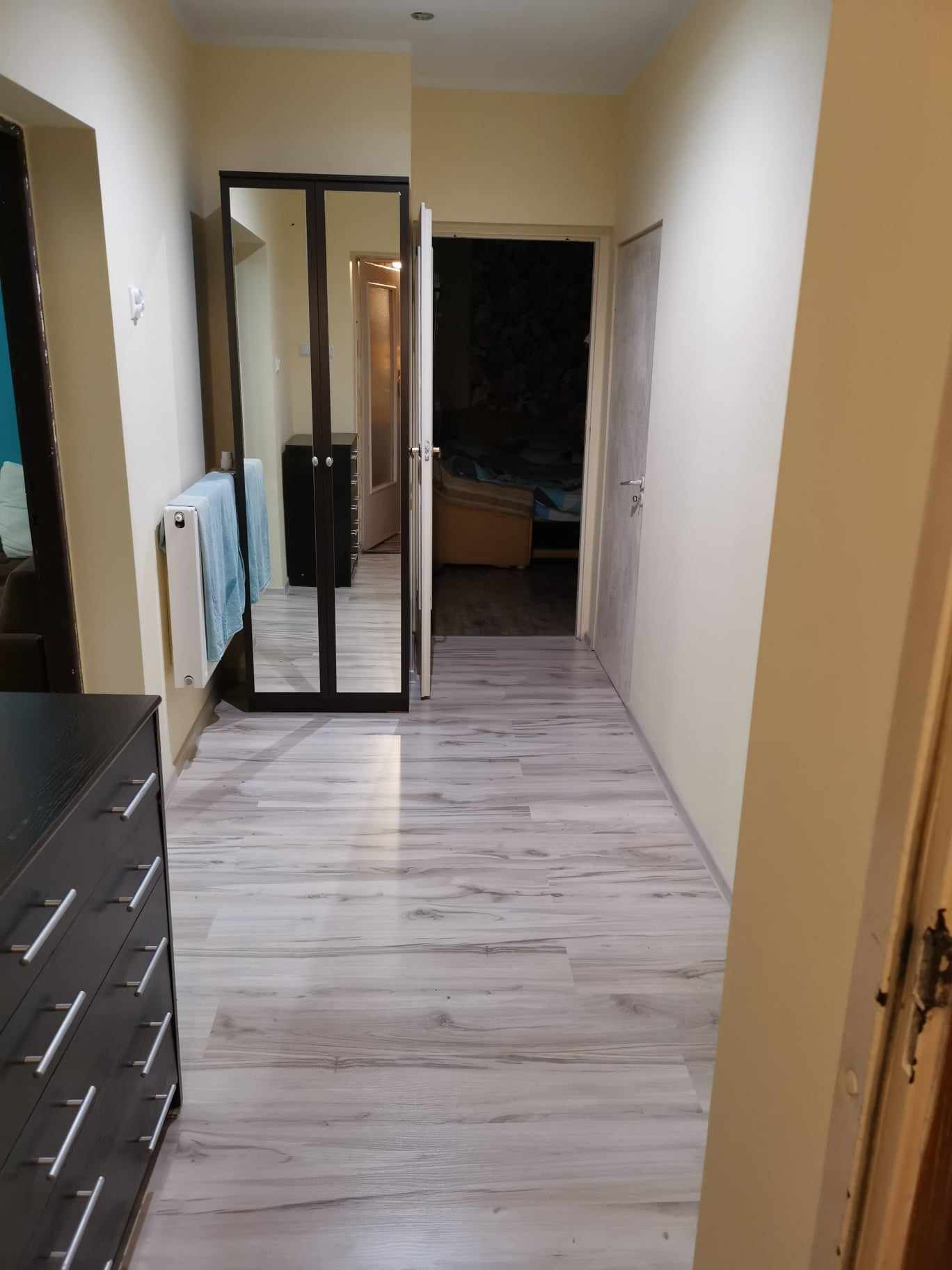 Wnętrze mieszkania z nową podłogą imitującą drewno, widoczna szafa z lustrem, grzejnik z ręcznikiem i fragment komody.