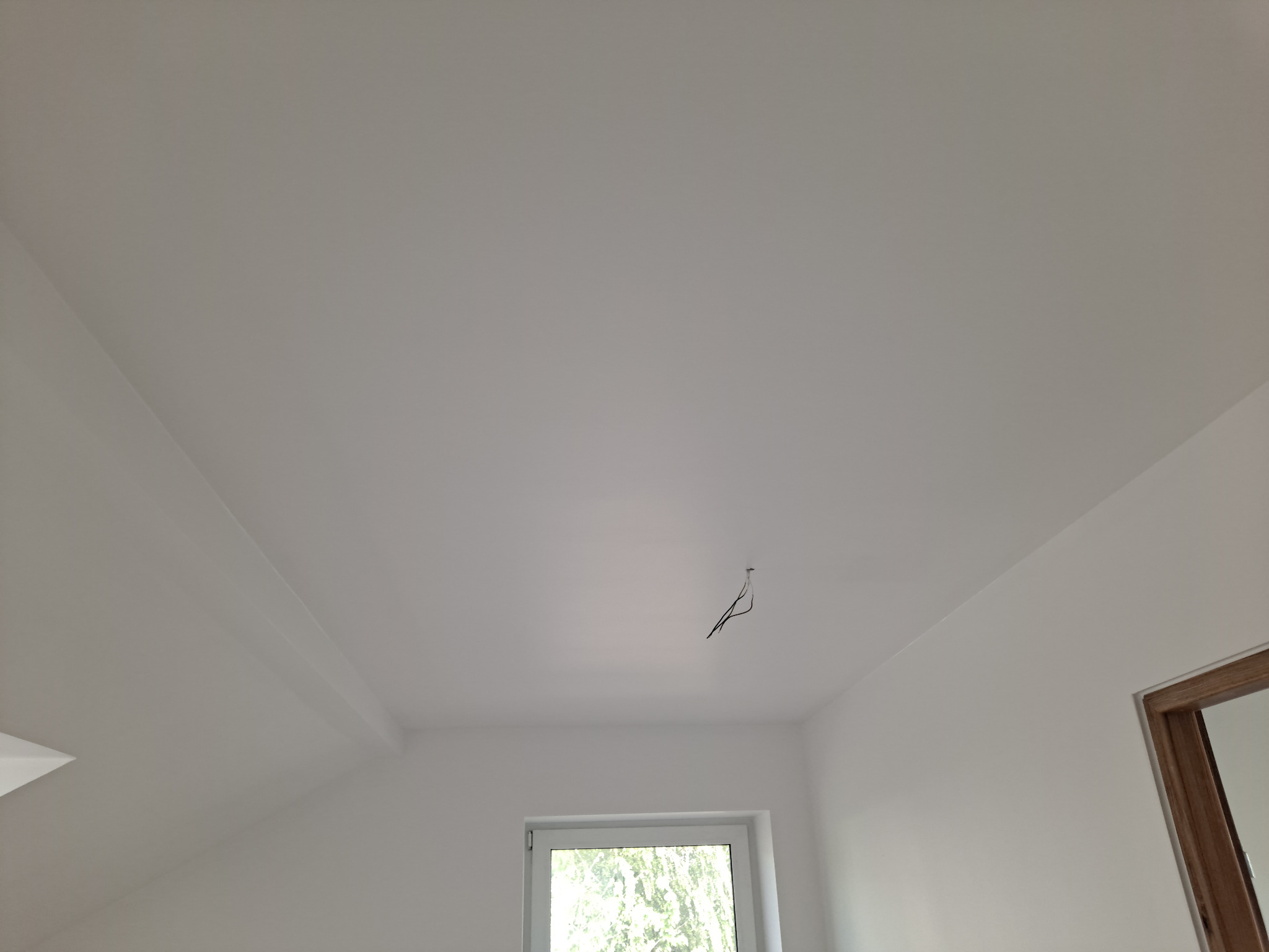 Białe, świeżo pomalowane wnętrze pokoju z oknem, widoczne wystające kable ze ściany pod sufitem, sugerujące przygotowanie do montażu oświetlenia.